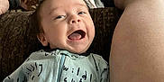 Thomas is registered to the contest to win money with this photo: baby, infant, smiling, happy, onesie, clothing, person, face, leg, footwear, sandal, indoor, table, snacks, plastic_bag, hair, skin, joy, expression, couch
