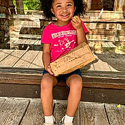Ma'Leah joined the competition — help win amazing prizes! child, girl, smiling, curly_hair, pink_bow, pink_tshirt, denim_shorts, sitting, wooden_floor, snack, paper_bag, white_socks, casual_shoes, outdoor, sunny, reflection, glass_door, wooden_background, happy, portrait