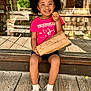 child, girl, smiling, curly_hair, pink_bow, pink_tshirt, denim_shorts, sitting, wooden_floor, snack, paper_bag, white_socks, casual_shoes, outdoor, sunny, reflection, glass_door, wooden_background, happy, portrait