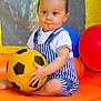 Milo participe au concours pour gagner de l'argent avec cette photo : toddler, child, soccer_ball, yellow_ball, striped_overalls, white_shirt, inflatable, orange, red_ball, blue_ball, play_area, mesh_background, cute, sitting, outdoor, playtime, young_child, baby, holding, fun