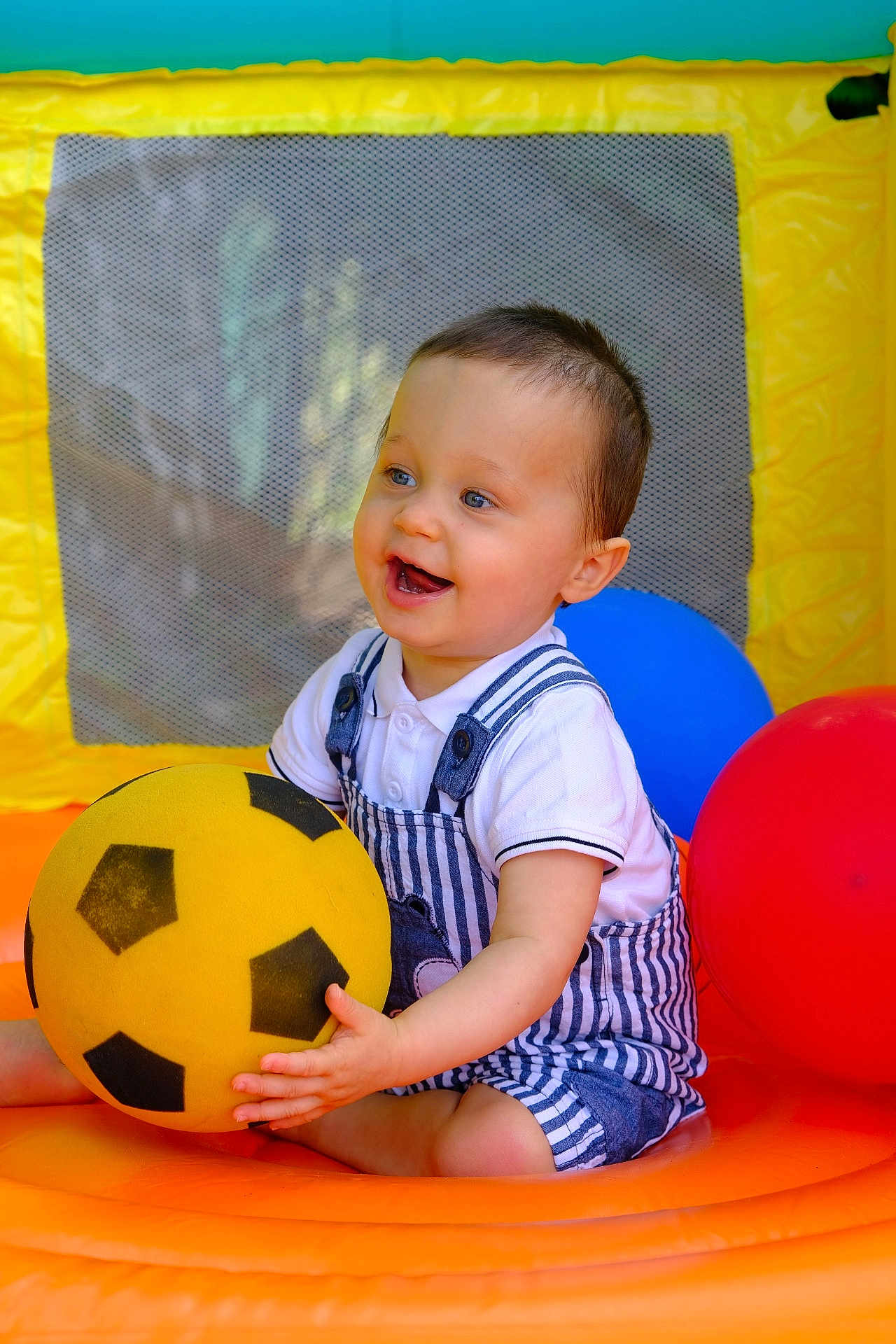 Milo a rejoint le concours — aidez-le/la à gagner de superbes lots ! toddler, child, smiling, ball, soccer_ball, inflatable, play_area, yellow, red, blue, mesh, happy, sitting, clothing, overalls, shorts, polo_shirt, indoor, toy, cute