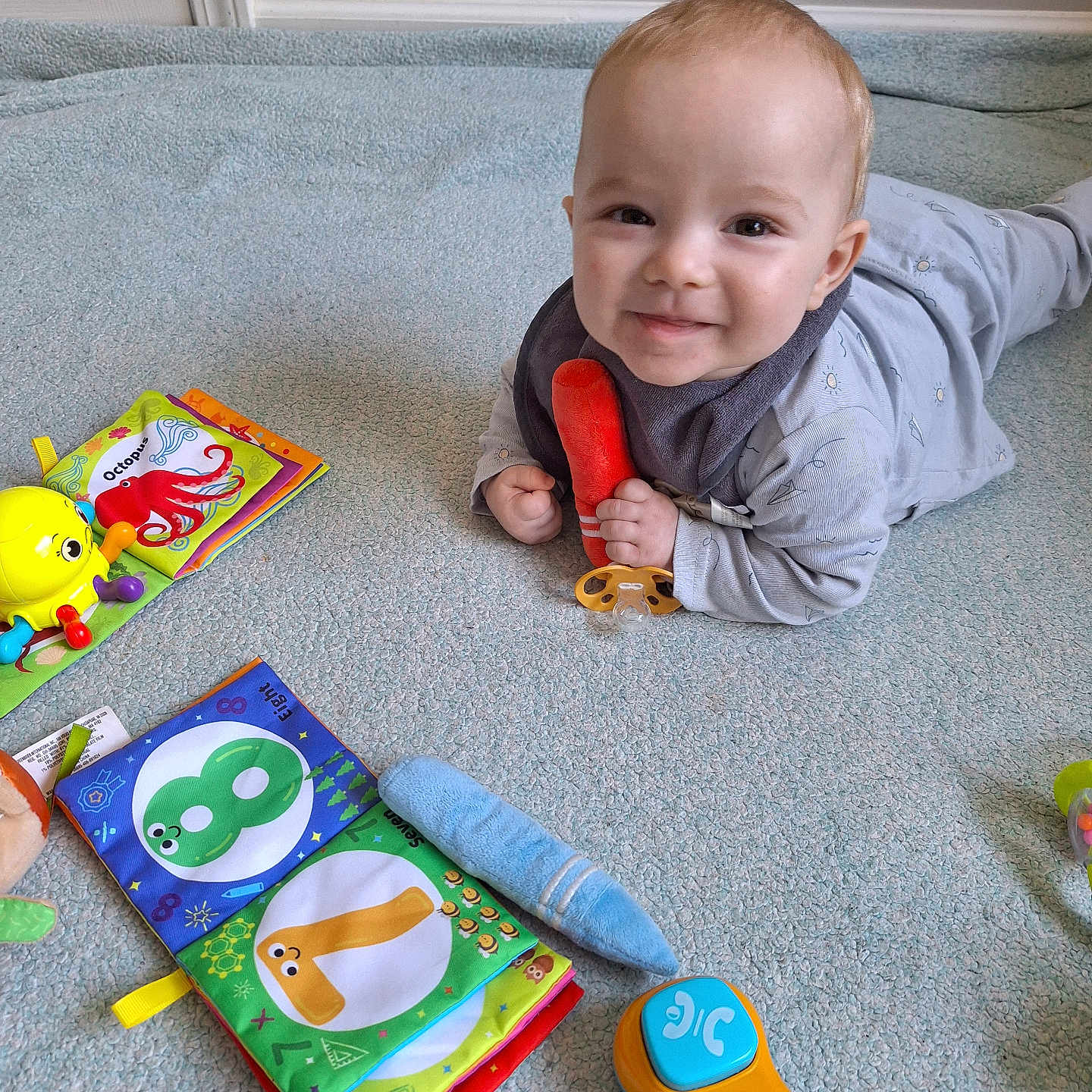 Jayce joined the competition — help win amazing prizes! baby, blue_carpet, carpet, child, clothing, colorful, cute, fabric_book, floor, happy, indoor, infant, lying_down, orange_toy, pacifier, playthings, playtime, smiling, toy, young_child