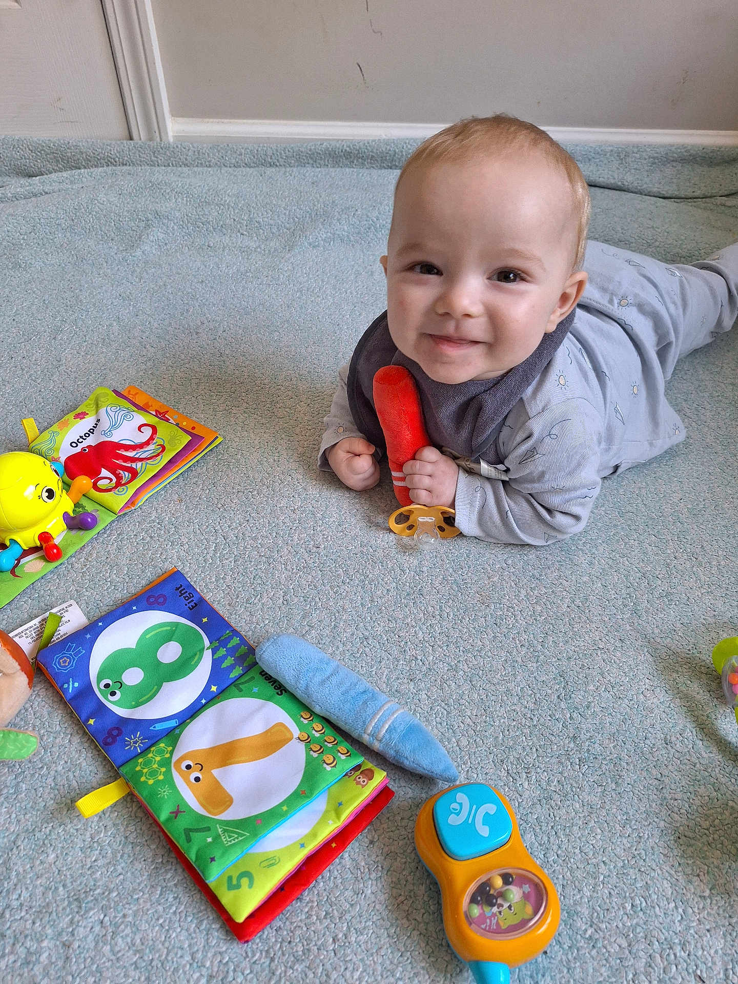 Jayce joined the competition — help win amazing prizes! baby, child, smiling, toy, carpet, fabric_book, orange_toy, blue_carpet, indoor, playtime, pacifier, clothing, infant, cute, colorful, floor, lying_down, happy, young_child, playthings
