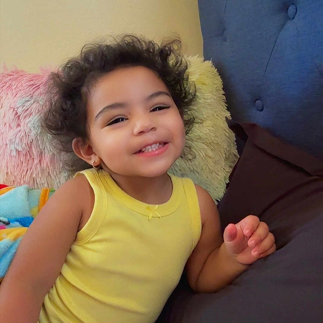 Jazminñ joined the competition — help win amazing prizes! child, toddler, smiling, curly_hair, yellow_tank_top, bed, pillow, smartphone, indoor, happy, casual_clothing, colorful, couch, furniture, person, portrait, relaxed, technology, cute, home