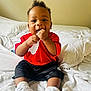 Nhyree is registered to the contest to win money with this photo: baby, bed, black_shorts, casual_clothing, chewing, child, curly_hair, cute, footwear, happy, indoor, infant, person, playing, red_shirt, sitting, smiling, white_bedding, white_sneakers, young_child