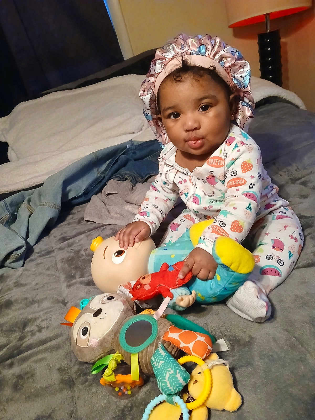 Dior is registered to the contest to win money with this photo: baby, child, pajamas, bonnet, toy, stuffed_animal, bed, indoor, lamp, blanket, cute, infant, soft_toy, sitting, playful, person, face, expression, cozy, colorful