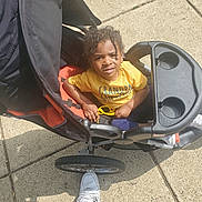 Derek is registered to the contest to win money with this photo: toddler, child, stroller, yellow_shirt, sunglasses, curly_hair, sidewalk, outdoor, sunlight, casual, person, shoes, concrete, black_stroller, orange_stroller, baby, seat, daytime, expression, portrait