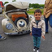 Lucas a rejoint le concours — aidez-le/la à gagner de superbes lots ! child, boy, vintage_car, volkswagen_beetle, open_hood, suitcase, spare_tire, smiling, jacket, sneakers, adult, outdoor, asphalt, cloudy_sky, greenery, people, parking_lot, license_plate, casual_clothing, happy