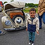 child, boy, vintage_car, volkswagen_beetle, open_hood, suitcase, spare_tire, smiling, jacket, sneakers, adult, outdoor, asphalt, cloudy_sky, greenery, people, parking_lot, license_plate, casual_clothing, happy