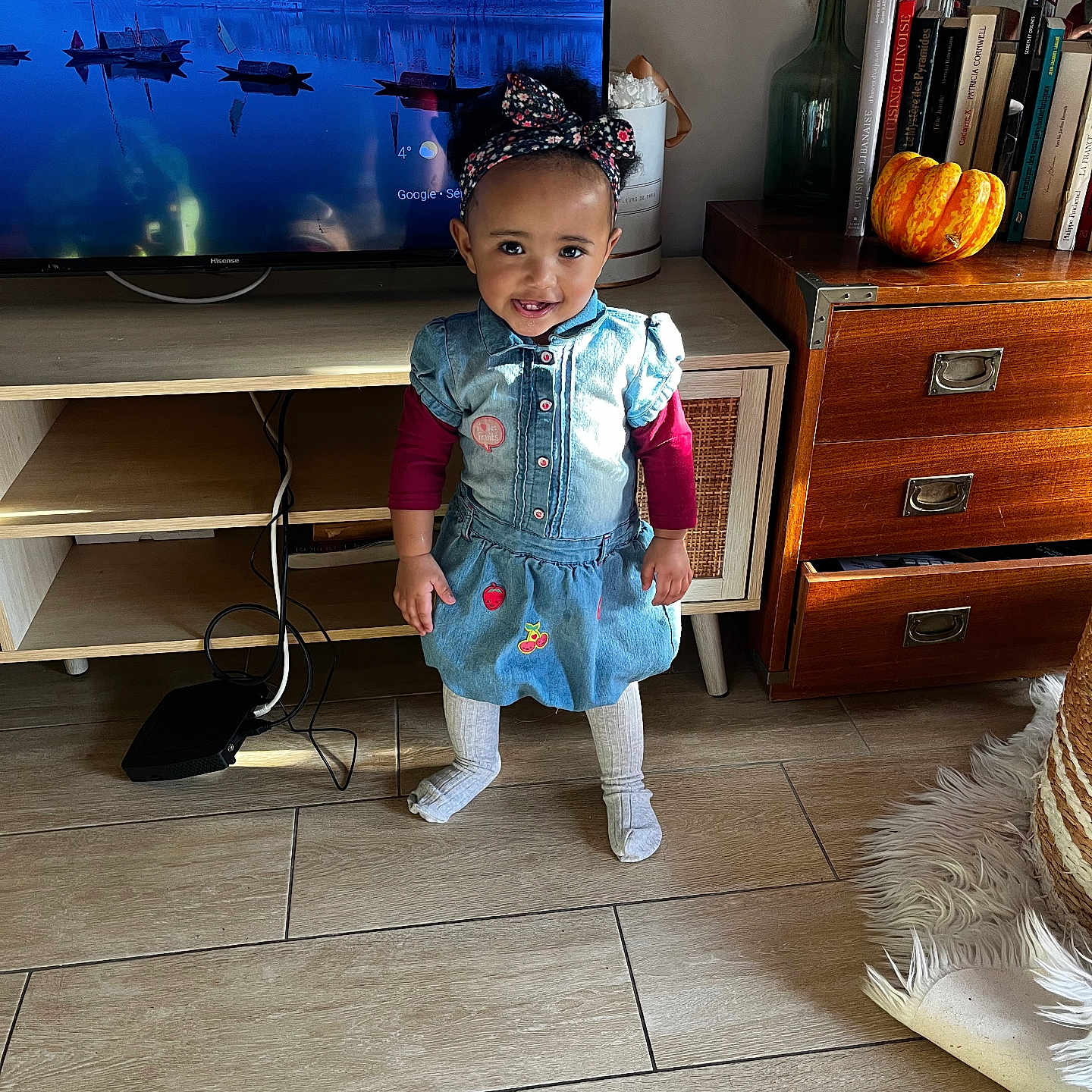 Seta a rejoint le concours — aidez-le/la à gagner de superbes lots ! books, cabinet, child, cozy, cute, decor, denim_dress, floor_tiles, happy, headband, indoor, living_room, person, portrait, pumpkin, smiling, standing, television, toddler, wooden_floor