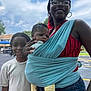 woman, baby, child, glasses, hoop_earrings, blue_wrap, red_shirt, yellow_pants, white_socks, smile, outdoor, sky, clouds, street, suburb, family, holding, person, portrait, daylight