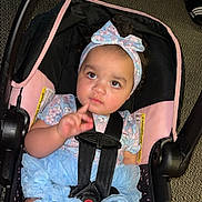 Makiera is registered to the contest to win money with this photo: baby, child, car_seat, headband, bow, floral_clothing, ruffled_shorts, straps, sitting, indoor, curious, infant, person, pink, black, blue, car_seat_strap, floor, footwear, expression