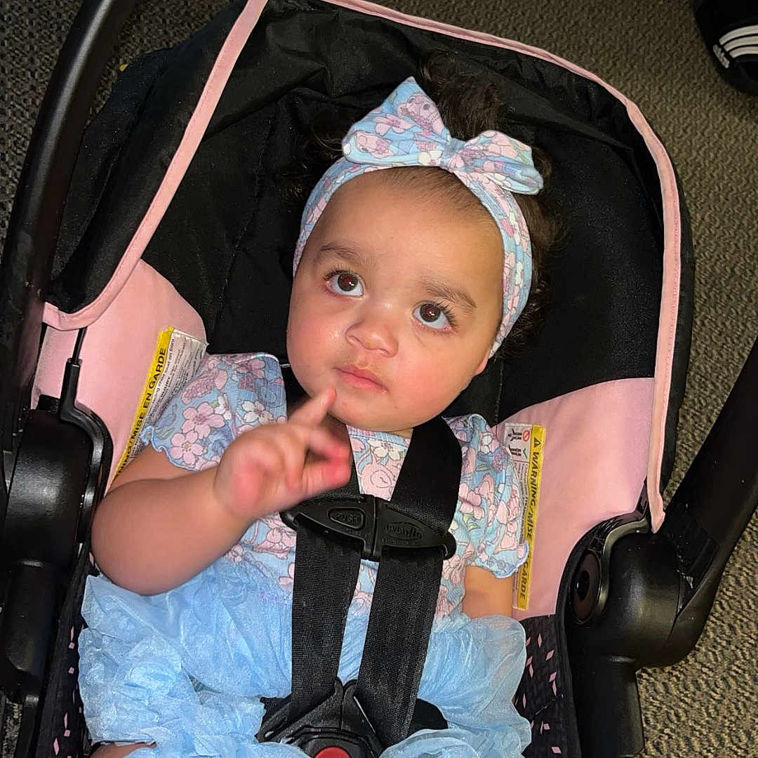 Makiera is registered to the contest to win money with this photo: baby, black, blue, bow, car_seat, car_seat_strap, child, curious, expression, floor, floral_clothing, footwear, headband, indoor, infant, person, pink, ruffled_shorts, sitting, straps