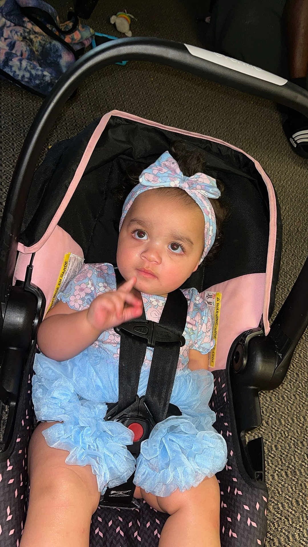 Makiera is registered to the contest to win money with this photo: baby, child, car_seat, headband, bow, floral_clothing, ruffled_shorts, straps, sitting, indoor, curious, infant, person, pink, black, blue, car_seat_strap, floor, footwear, expression