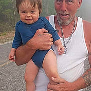 Memphis is registered to the contest to win money with this photo: man, baby, child, holding, outdoor, road, trees, fog, smile, tattoos, necklace, white_tank_top, blue_clothing, socks, casual, portrait, person, nature, daytime, happy