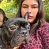 dog, french_bulldog, person, woman, portrait, selfie, outdoor, garden, trees, leaves, sunlight, red_harness, leash, animal, pet, face, ears, brown_hair, leopard_print, close_up