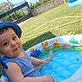 toddler, child, baby, pool, inflatable_pool, water, blue_hat, blue_tank_top, outdoor, grass, summer, sunny, playful, cute, happy, water_droplets, backyard, garden, fence, daytime