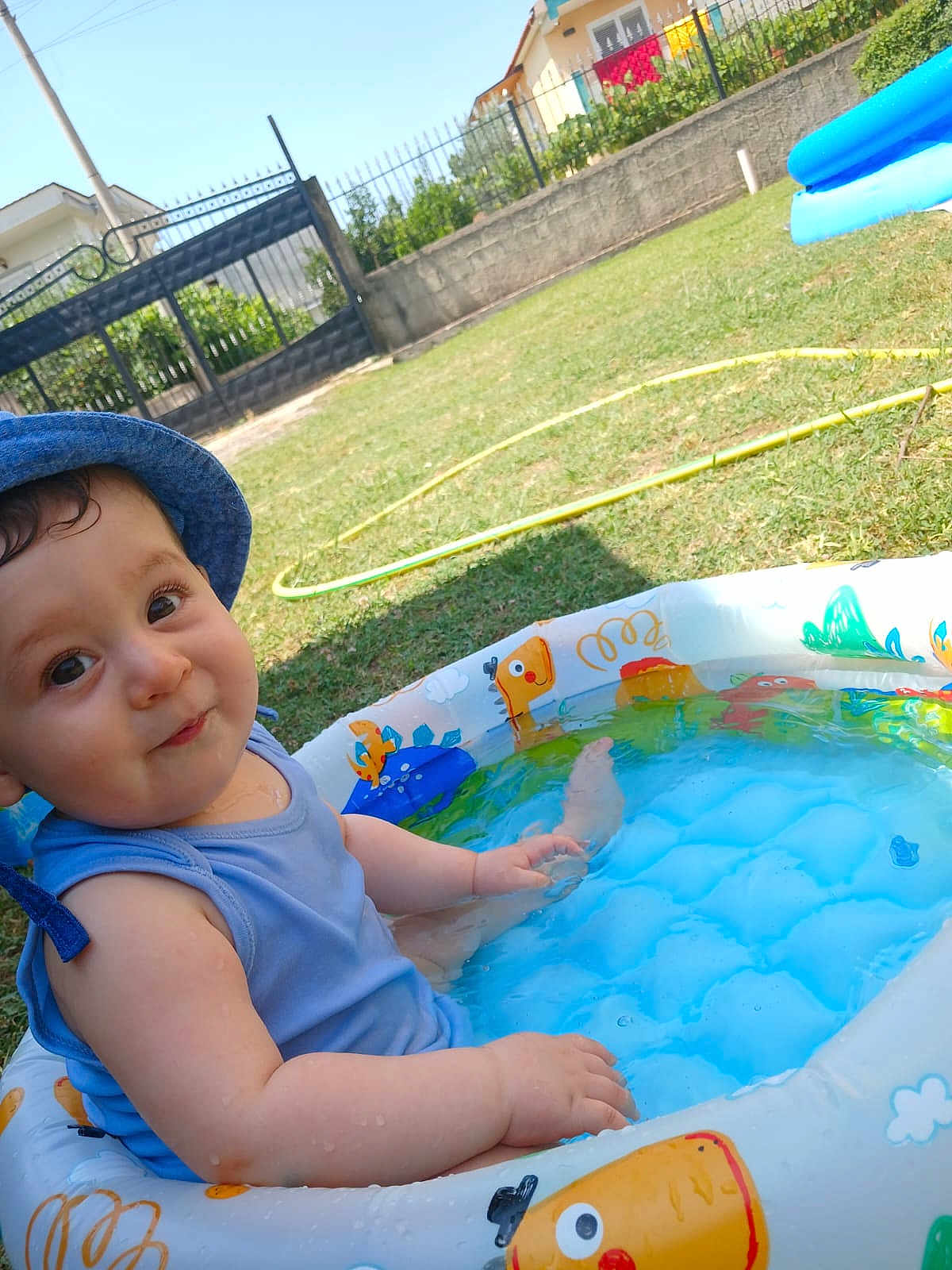 Amadeo joined the competition — help win amazing prizes! toddler, child, baby, pool, inflatable_pool, water, blue_hat, blue_tank_top, outdoor, grass, summer, sunny, playful, cute, happy, water_droplets, backyard, garden, fence, daytime