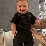 Nylann participe au concours pour gagner de l'argent avec cette photo : baby, black_clothing, chair, child, cushion, cute, happy, indoor, overalls, patterned_wallpaper, person, portrait, shoes, smiling, standing, supporting_hands, tattooed_arms, tongue_out, wall, young_child