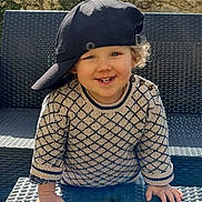 Gabriel a rejoint le concours — aidez-le/la à gagner de superbes lots ! child, toddler, smiling, hat, cap, sweater, patterned_clothing, table, outdoor, sunlight, shadow, stone_wall, greenery, happy, portrait, face, hands, curly_hair, bench, playful