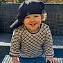 child, toddler, smiling, hat, cap, sweater, patterned_clothing, table, outdoor, sunlight, shadow, stone_wall, greenery, happy, portrait, face, hands, curly_hair, bench, playful