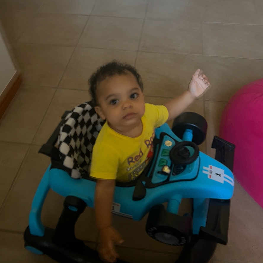 Jakaiven is registered to the contest to win money with this photo: toddler, child, baby, sitting, ride_on_toy, toy_vehicle, blue_toy, yellow_shirt, waving, curious, indoors, tile_floor, pink_ball, playroom, portrait, face, person, casual_clothing, home, daylight
