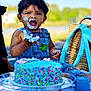 child, toddler, cake, blue_cake, frosting, outdoor, picnic, denim_overalls, smiling, face_paint, happy, celebration, food, birthday, grass, sunny, sitting, hand, baby, playful