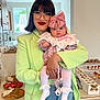 woman, baby, indoor, glasses, green_blazer, jeans, pink_bow, floral_dress, table, food, snacks, treats, holding, portrait, person, happy, face, headwear, cute, celebration