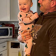 Alena is registered to the contest to win money with this photo: baby, man, kitchen, smile, laughing, holding, pajamas, reindeer, christmas, face, person, hoodie, bandana, cabinet, microwave, counter, cup, door, indoor, happy