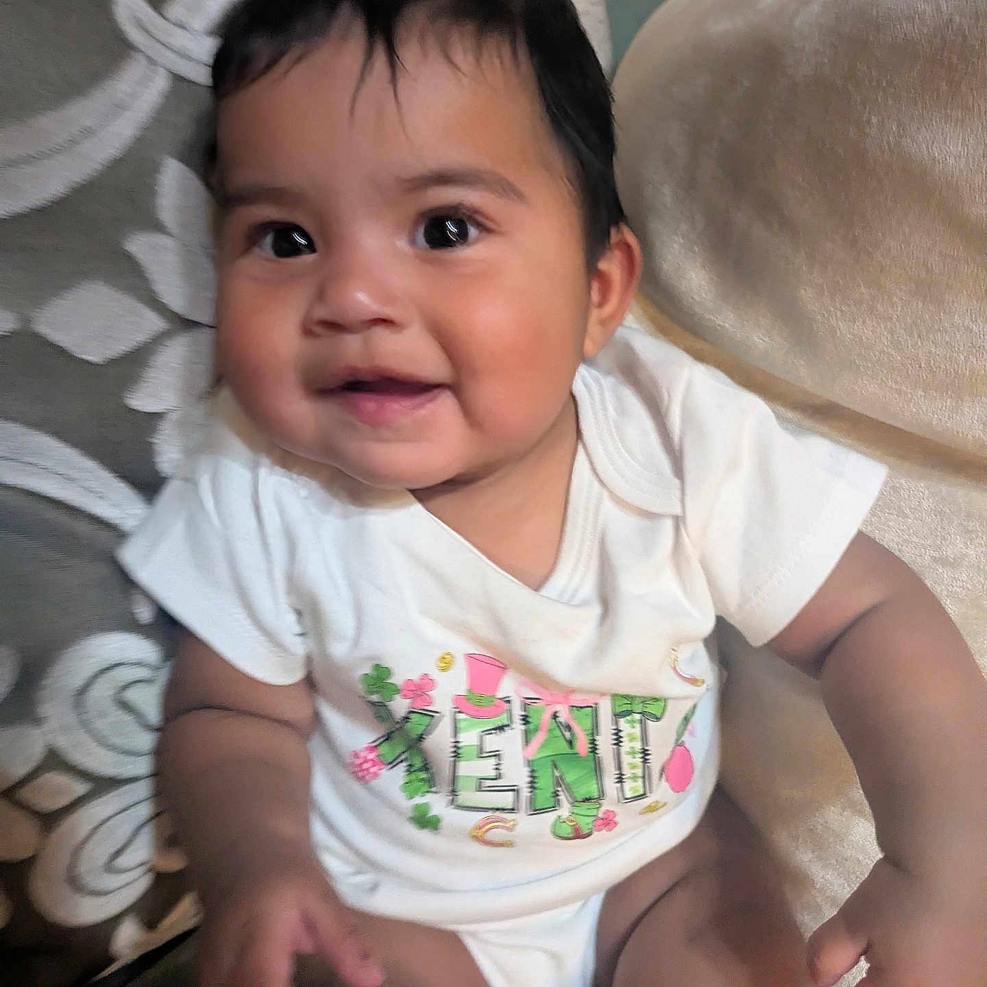 Xenia joined the competition — help win amazing prizes! adorable, baby, big_eyes, chubby_cheeks, closeup, clothing, couch, cushion, dark_hair, fabric, hand, happy, indoor, infant, living_room, onesie, pillow, portrait, sitting, smiling