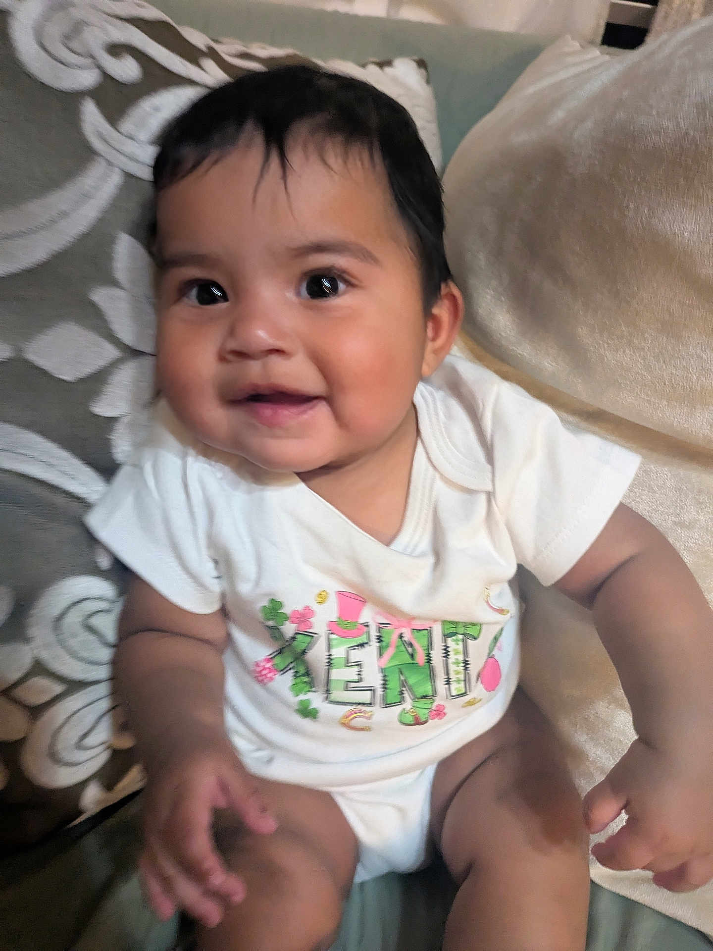 Xenia joined the competition — help win amazing prizes! adorable, baby, big_eyes, chubby_cheeks, closeup, clothing, couch, cushion, dark_hair, fabric, hand, happy, indoor, infant, living_room, onesie, pillow, portrait, sitting, smiling