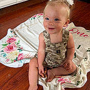 Daisy is registered to the contest to win money with this photo: toddler, child, baby, smiling, sitting, blanket, floral, hair_bows, overalls, camouflage, floor, wooden_floor, indoors, cute, happy, person, portrait, skin, face, blue_eyes
