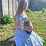 girl, cat, grass, garden, sunlight, dress, blue_dress, outdoor, fence, greenery, pet, animal, young, long_hair, barefoot, fluffy_cat, tabby_cat, sitting, holding, peaceful