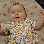 Leah is registered to the contest to win money with this photo: baby, child, infant, smile, face, floral_dress, blanket, pattern, happy, cute, person, young_child, lying_down, indoor, skin, head, hair, clothing, baby_girl, motion_blur