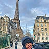 toddler, child, smiling, winter_clothing, beanie, outdoor, eiffel_tower, paris, cityscape, street, sidewalk, buildings, people, urban, daytime, cloudy_sky, happy, portrait, tourist, landmark