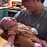 Nova joined the competition — help win amazing prizes! baby, young_man, holding, kitchen, cap, chain_necklace, onesie, mittens, wood_floor, stove, cabinets, smiling, tattoo, watch, appliance, indoor, person, face, happy, love