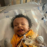 Lafenis Owens joined the competition — help win amazing prizes! baby, smiling, hospital, crib, medical_tubes, yellow_shirt, mittens, infant, face, bed, healthcare, child, cute, lying_down, indoors, person, happy, medical_equipment, newborn, cozy