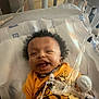 baby, smiling, hospital, crib, medical_tubes, yellow_shirt, mittens, infant, face, bed, healthcare, child, cute, lying_down, indoors, person, happy, medical_equipment, newborn, cozy