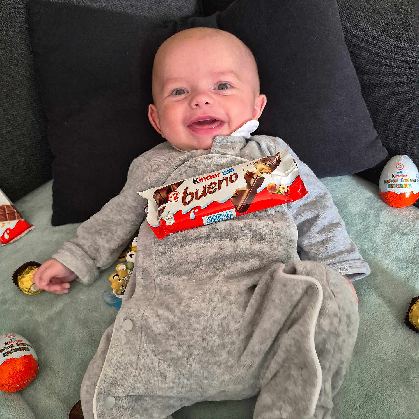 Aylann a rejoint le concours — aidez-le/la à gagner de superbes lots ! baby, smiling, infant, onesie, gray_clothing, pillow, couch, blanket, chocolate, kinder_bueno, kinder_egg, ferrero_rocher, food, sweet, snack, happy, indoors, person, head, face