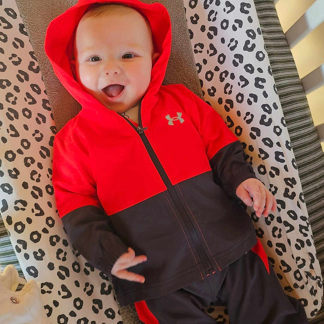 Isaac a rejoint le concours — aidez-le/la à gagner de superbes lots ! baby, child, hoodie, red_clothing, black_clothing, smiling, tongue_out, changing_pad, animal_print, striped_socks, striped_mat, indoor, infant, cute, playful, person, apparel, fabric, happy, lying_down