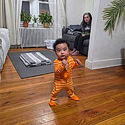 Aaron is registered to the contest to win money with this photo: toddler, child, baby, standing, orange_onesie, striped_clothing, smiling, indoor, living_room, wooden_floor, adult, woman, houseplants, radiator, window, curtains, sofa, laundry_basket, blanket, portrait