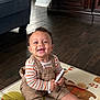 baby, child, smiling, curly_hair, overalls, striped_shirt, play_mat, remote_control, hairbrush, wooden_floor, couch, cabinet, indoor, happy, sitting, person, furniture, floor, toy, home