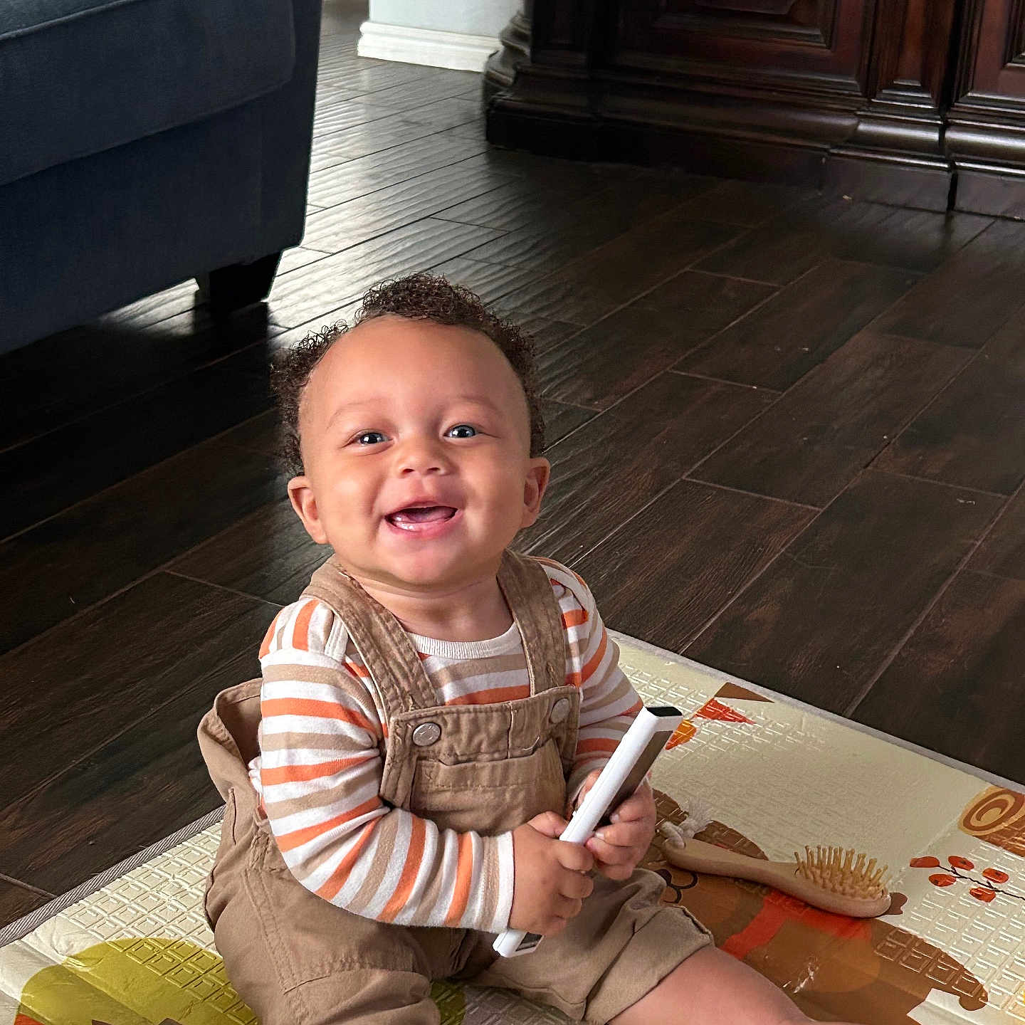 Jahbari joined the competition — help win amazing prizes! baby, cabinet, child, couch, curly_hair, floor, furniture, hairbrush, happy, home, indoor, overalls, person, play_mat, remote_control, sitting, smiling, striped_shirt, toy, wooden_floor