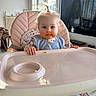baby, high_chair, messy_face, blue_eyes, pony_tail, indoor, window, television, reflection, pastel_colors, clothing, bib, hands, footwear, floor, furniture, curious, child, home, portrait