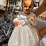 toddler, child, dress, white, lace, cardigan, outdoor, sunlight, smile, hair, person, footwear, stone, adult_arm, holding, bread, portrait, happy, young_child, fashion