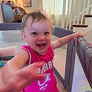 Lola is registered to the contest to win money with this photo: toddler, child, smiling, pink_clothing, playpen, indoor, carpet, curtains, stairs, lamp, furniture, happy, person, home, living_room, toy, barefoot, face, gesture, young_child