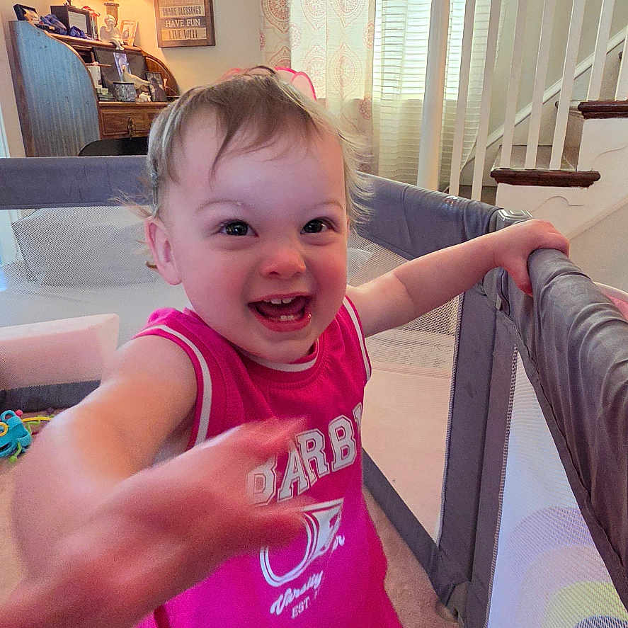 Lola is registered to the contest to win money with this photo: toddler, child, smiling, pink_clothing, playpen, indoor, carpet, curtains, stairs, lamp, furniture, happy, person, home, living_room, toy, barefoot, face, gesture, young_child