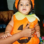 Aariv is registered to the contest to win money with this photo: baby, child, pumpkin_costume, halloween, orange_hat, costume, portrait, smiling, indoor, adult_hand, supportive_hold, toy, playroom, sitting, infant, colorful_clothing, home, face, shorts, seasonal