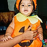 baby, child, pumpkin_costume, halloween, orange_hat, costume, portrait, smiling, indoor, adult_hand, supportive_hold, toy, playroom, sitting, infant, colorful_clothing, home, face, shorts, seasonal