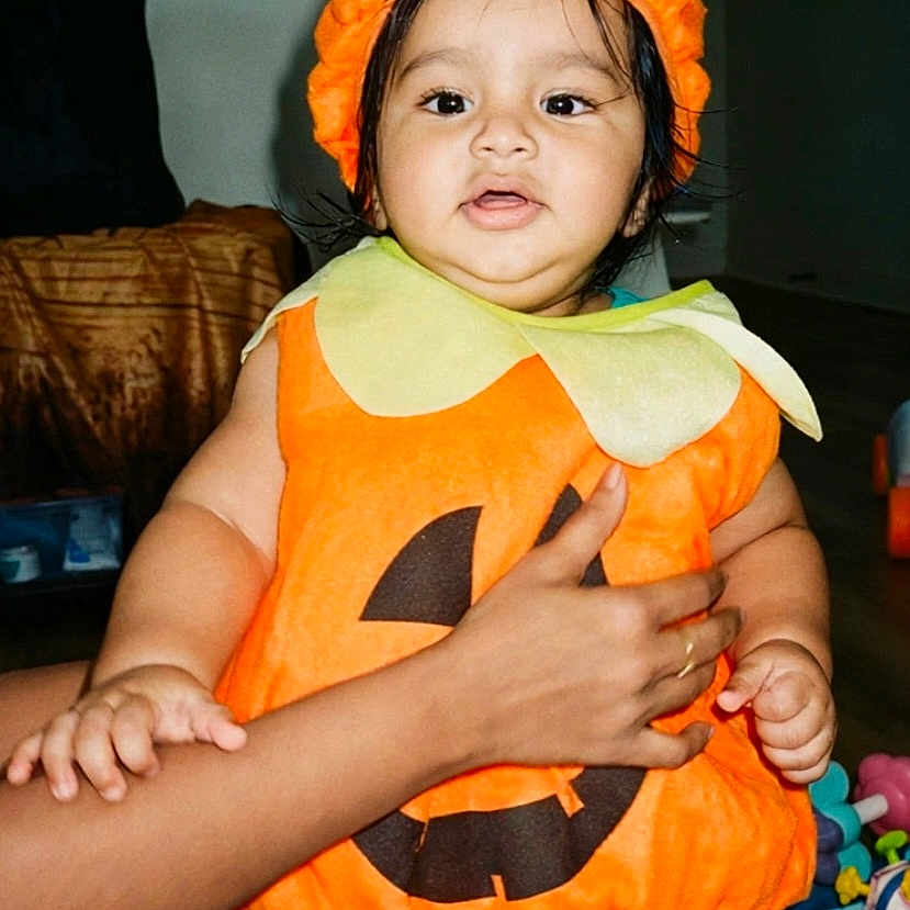 Aariv is registered to the contest to win money with this photo: baby, child, pumpkin_costume, halloween, orange_hat, costume, portrait, smiling, indoor, adult_hand, supportive_hold, toy, playroom, sitting, infant, colorful_clothing, home, face, shorts, seasonal